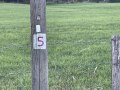 A wooden post with the number 5 stands on a green meadow, surrounded by a wire fence., © André Szybalsky