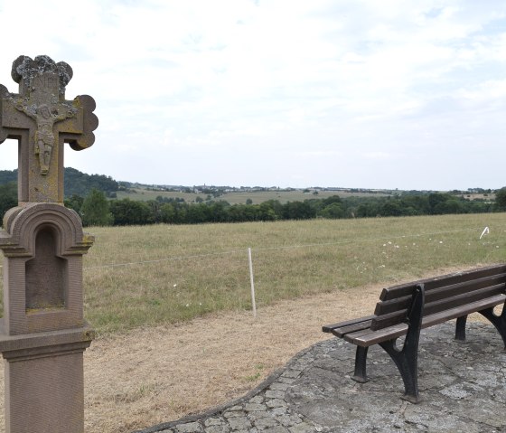 Bank en stenen kruis in een landelijk landschap met weids uitzicht over velden en heuvels., © TI Bitburger Land