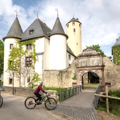 Two cyclists ride past the historic Rittersdorf Castle, surrounded by green trees and a clear sky., &copy; Eifel-Tourismus GmbH/Dominik Ketz