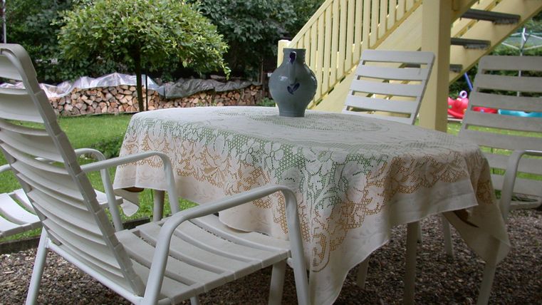 A cozy table with a sitting surface made of white wood and a decorative tablecloth. In the background, a green tree and a staircase are visible.