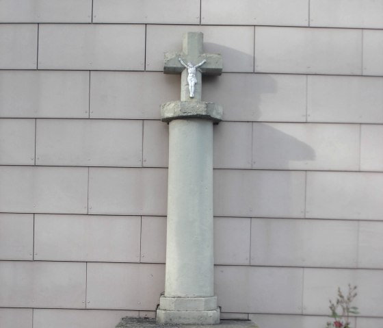 A stone wayside cross with a figure of Jesus stands in front of a house wall. A small wooden fence and plants are visible in the foreground., © Conny Meier
