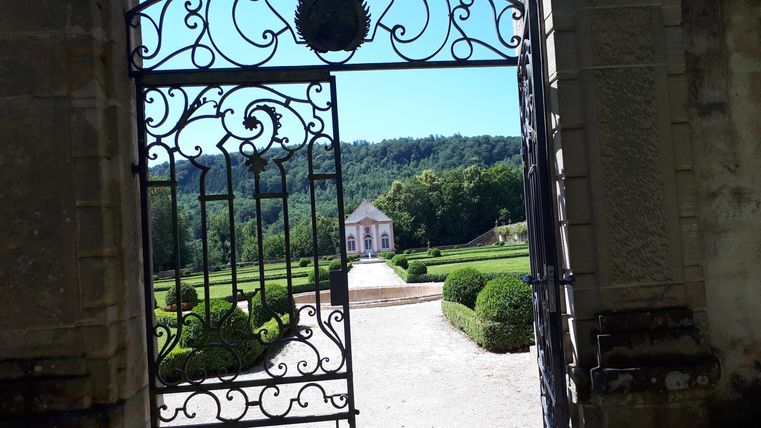 Ein schöner Blick durch ein schmiedeeisernes Tor auf einen gepflegten Garten und ein charmantes Gebäude im Hintergrund. Grüne Bäume und blauer Himmel ergänzen die Landschaft.