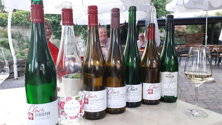 Eine Reihe von Weinflaschen steht auf einem Tisch, einige sind mit rotem Korken und andere mit grünem Korken. Im Hintergrund sind verschwommene Personen und eine gemütliche Atmosphäre.