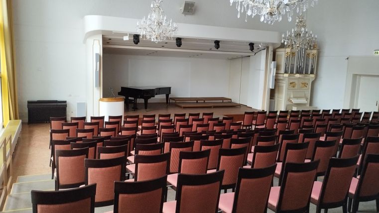 Een grote evenementenhal met veel rode stoelen. Op het podium staat een piano en op de achtergrond staat een mooie orgel.