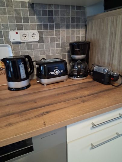 Eine moderne Küchentheke mit einem Wasserkocher, einem Toaster, einer Kaffeemaschine und einer Küchenmaschine. Die Geräte sind in Schwarz und Edelstahl gehalten und stehen auf einer Holzoberfläche.