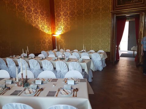 An elegant banquet hall with festively laid tables and chairs in white. The walls are decorated with golden patterns, and soft lighting creates an inviting atmosphere.