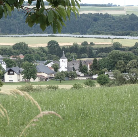 Ein Dorf mit einer Kirche in der Mitte, umgeben von grünen Feldern und Bäumen, unter einem bewölkten Himmel., © TI Bitburger Land