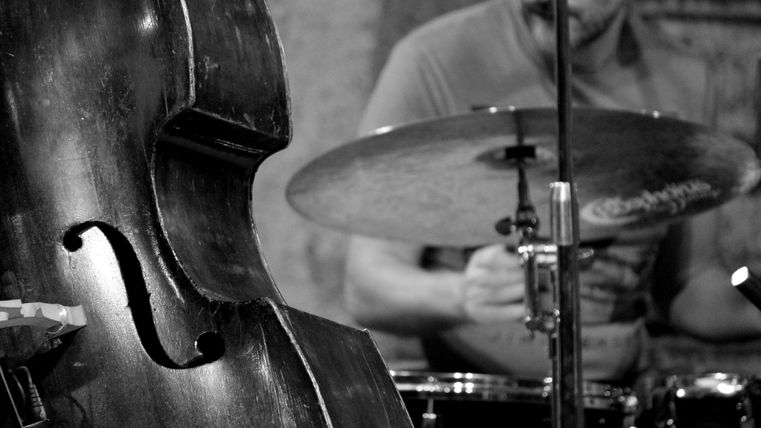 A double bass is in the foreground, while a drummer is visible in the background. The scene conveys a musical atmosphere.