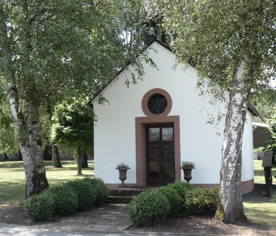 A small white chapel with brown windows, surrounded by trees and shrubs, stands in a green landscape., © TI Bitburger Land