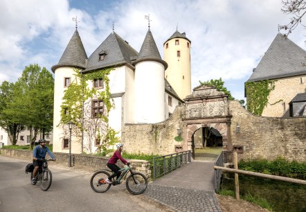 Deux cyclistes passent devant le château historique de Rittersdorf, entouré d'arbres verts et d'un ciel dégagé., © Eifel-Tourismus GmbH/Dominik Ketz