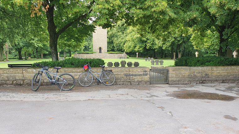 Zwei Fahrräder vor einer Steinmauer mit Friedhof im Hintergrund.