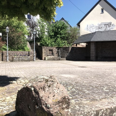 Ein ruhiger Dorfplatz mit gepflastertem Boden, umgeben von Bäumen und einem Gebäude mit Wandmalerei. Laternen und Pflanzen schmücken den Platz., © Tourist-Information Bitburger Land