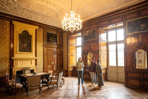 An elegant room with wooden walls and a large chandelier. Three people are standing at the table and looking at the furnishings.