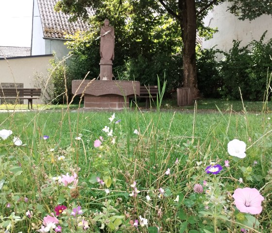 Une statue de Donatus se trouve dans un jardin. Des fleurs multicolores s'épanouissent au premier plan. Des bancs et un arbre sont visibles à l'arrière-plan., © TI Bitburger Land