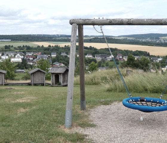 Playground with a blue swing and wooden huts, surrounded by green fields and a village in the background., © TI Bitburger Land