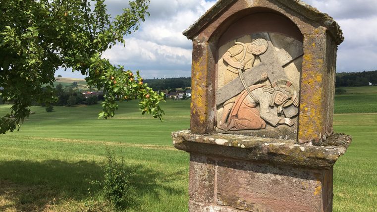 Steinrelief eines Kreuzwegs in ländlicher Landschaft mit Apfelbaum im Vordergrund.