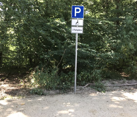 Ein Parkplatzschild für Behinderte im Wald. Es zeigt zwei verfügbare Parkplätze an. Umgeben von dichtem Grün und Bäumen., © TI Bitburger Land