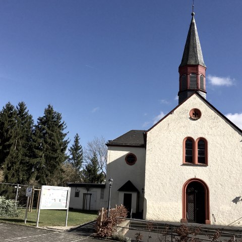 Eine Kirche in Wißmannsdorf mit spitzem Turm und roten Fensterrahmen vor einem klaren blauen Himmel., © TI Bitburger Land