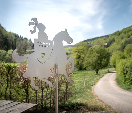 Metalen figuur van een ridder op een paard met een lans, voor een groen landschap en een pad. Op de achtergrond zijn bomen en een blauwe lucht te zien., © Tourist-Information Bitburger Land_Monika Mayer