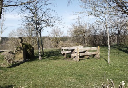 Rastplatz mit Holzbänken und steinernem Kreuz auf einer Wiese, umgeben von kahlen Bäumen unter blauem Himmel., © M. Bach