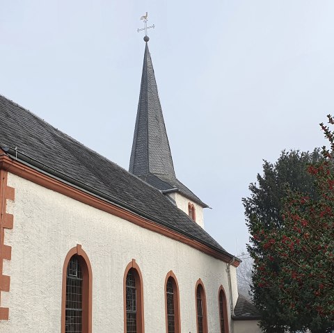 Eine Kirche in Idesheim mit einem Kreuz an der Fassade und einem Baum mit roten Beeren im Vordergrund., © TI Bitburger Land