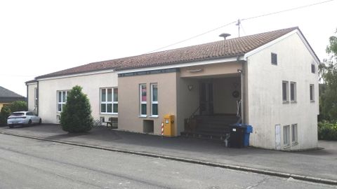 Ein einfaches Gebäude mit einem flachen Dach und mehreren Fenstern. Davor stehen ein Auto und einige Pflanzen.