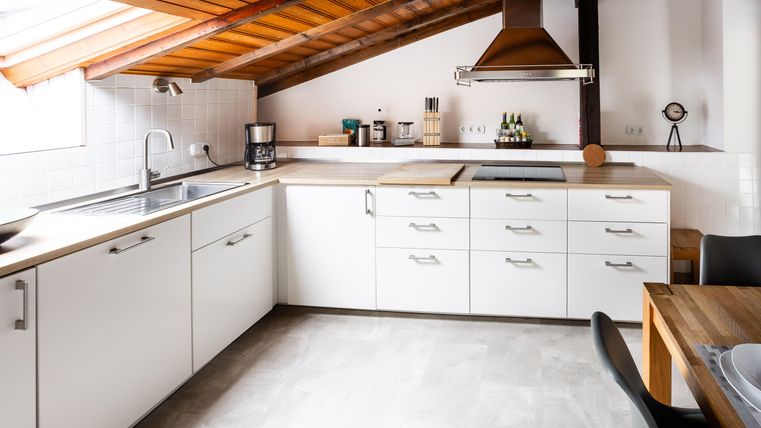 Eine moderne Küche mit weißen Schränken und Holzoberflächen. Das Dach ist schräg, und es gibt große Fenster, die viel Licht hereinlassen.