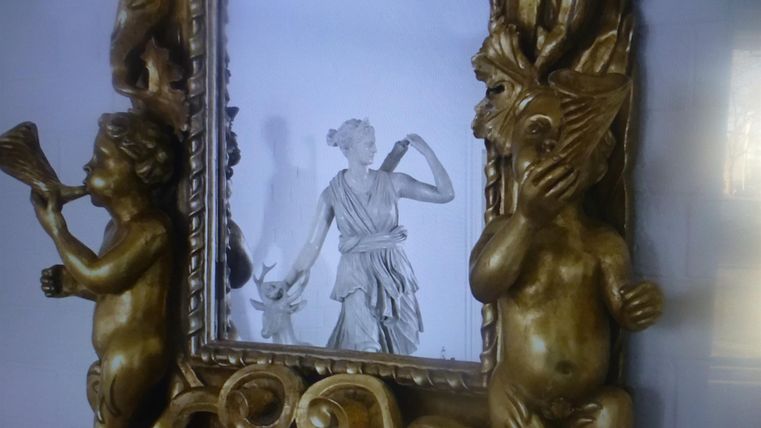 Ein kunstvoller Spiegelrahmen mit skulpturalen Details an den Seiten. In der Mitte ist eine Statue einer Frau, die ein Horn hält.