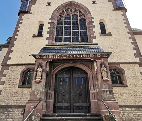 Eingang der Pfarrkirche Maria Königin in Dudeldorf mit steinerner Treppe, verzierten Holztüren und Statuen. Gotische Fenster und Ziegelsteindetails., © Melanie Lonien