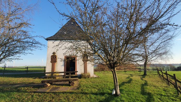 Small chapel on a meadow with trees and wind turbines in the background.