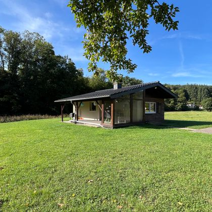 Ein gemütliches Ferienhaus auf einer grünen Wiese, umgeben von Bäumen. Der Himmel ist blau mit wenigen Wolken.