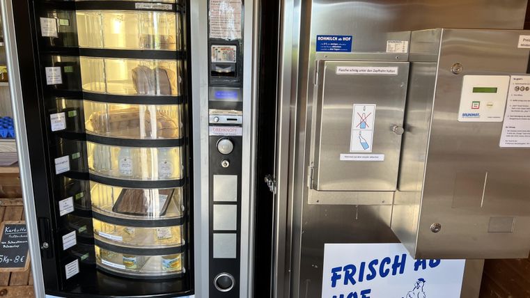 Ein Verkaufsautomat mit Snacks und Getränken steht neben einem Kühlschrank, der frische Produkte anbietet. Auf dem Kühlschrank befindet sich ein Schild mit der Aufschrift „FRISCH AB HOF“.