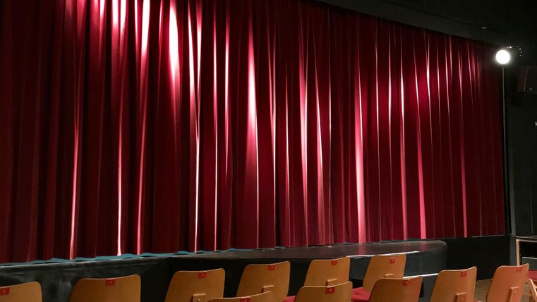 Ein Theater mit roten Vorhängen und einer Reihe von Stühlen vor der Bühne. Die Atmosphäre ist gespannt und bereit für eine Vorstellung.