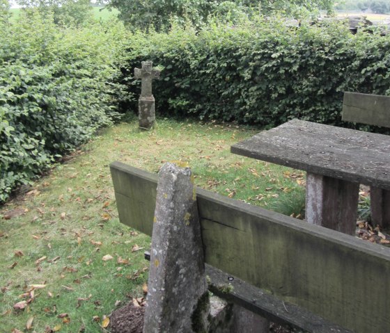 Ein Steinkreuz steht auf einer Wiese neben einem verwitterten Holztisch, umgeben von dichten Hecken und Bäumen., © M. Bach