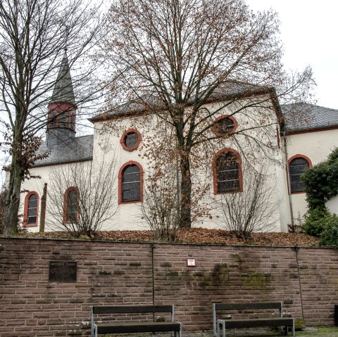 Die Kirche in Wißmannsdorf mit roten Fensterrahmen, umgeben von kahlen Bäumen und einer Steinmauer im Vordergrund., © TI Bitburger Land