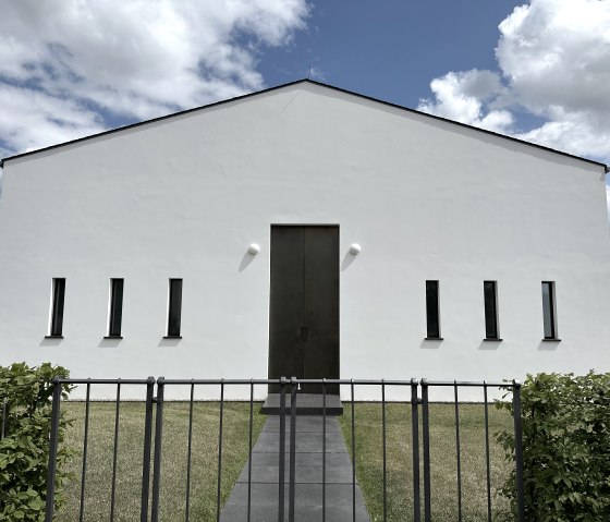Modern white building with narrow windows and large wooden gate, surrounded by lawn and fence, under a blue sky with clouds., © Felsenland Südeifel, Anna Carina Krebs