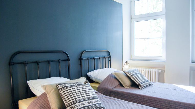 Ein helles Schlafzimmer mit zwei Einzelbetten und dekorativen Kissen. Das Zimmer hat große Fenster, die für viel Tageslicht sorgen.