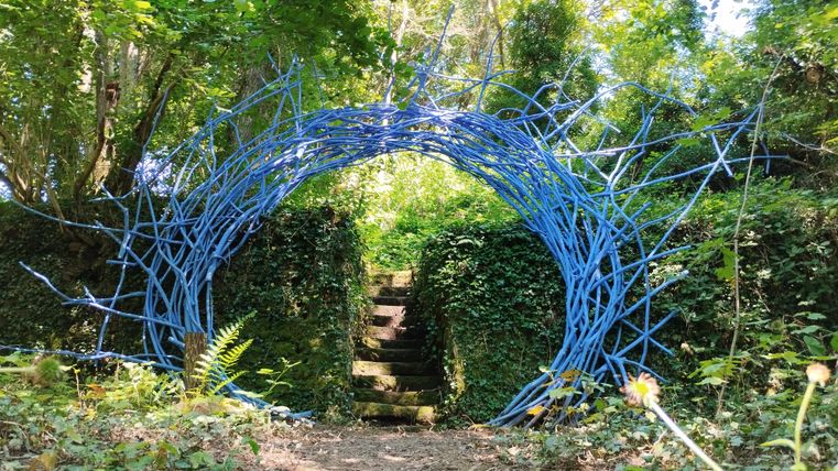 Une arche colorée faite de branches bleues mène à un jardin vert. Le chemin est entouré de plantes et d'arbres.