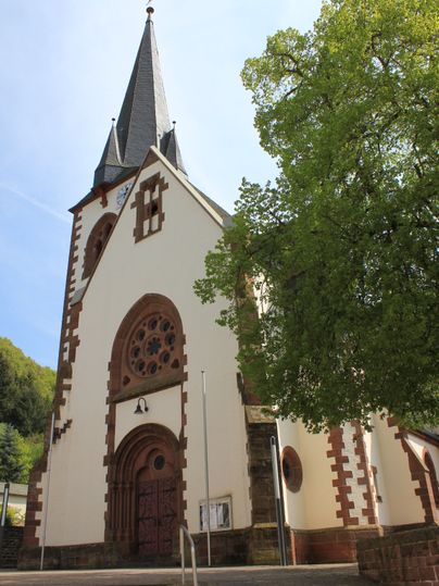 Kirche St. Quirinus in Malberg mit spitzem Turm und grünem Baum im Vordergrund.
