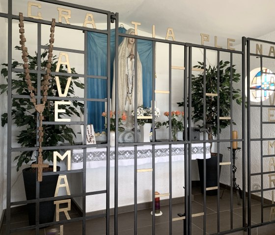 Chapelle avec statue de la Vierge Marie derrière une grille avec 'AVE MARIA'. Décorée de fleurs, de bougies et de chapelets., © Benjamin Milbach