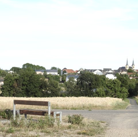 Eine Bank und ein Kreuz stehen vor einem Feld. Im Hintergrund ist ein Dorf mit einer Kirche und zwei Türmen zu sehen., © Thomas Neises