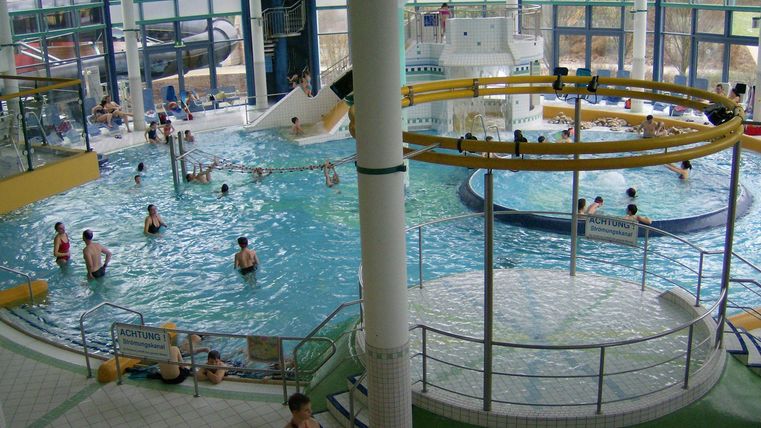 A large swimming pool with various water areas and many visitors. In the background, slides and large windows can be seen.