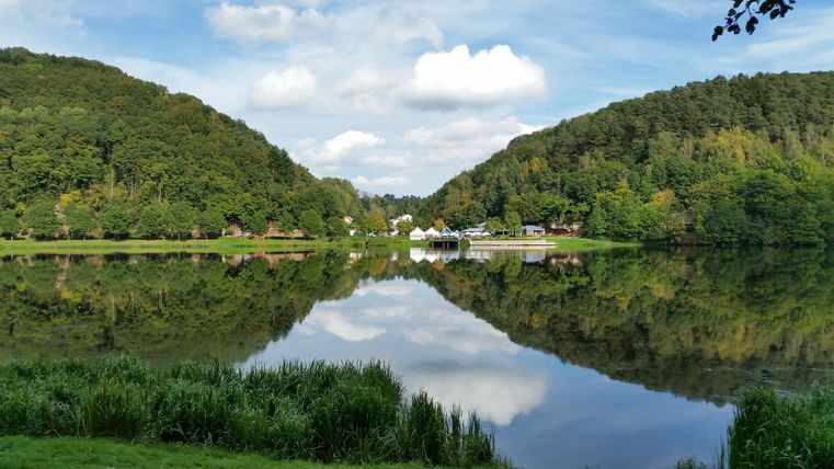 Ein ruhiger See, umgeben von bewaldeten Hügeln und grünem Gras. Der Himmel ist klar mit einigen Wolken, die sich im Wasser spiegeln.