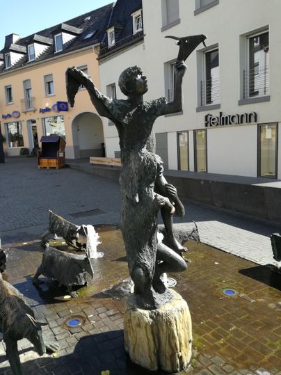 Eine Statue eines Jungen, der einen Papierflieger in die Höhe hält, steht in einer Stadtstraße. Um ihn herum sind weitere Skulpturen und ein ruhiger Brunnen.