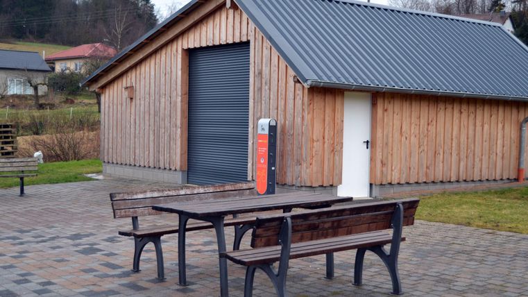 Un bâtiment en bois avec un grand rideau roulant et une porte blanche. Devant le bâtiment, il y a deux tables en bois et des bancs sur une zone pavée.