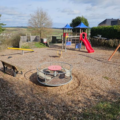 Ein Spielplatz mit einer Rutsche, einem Klettergerüst und einer Drehplattform. Der Boden ist mit Holzspänen bedeckt und es gibt einige Bäume und einen Ausblick auf die Landschaft.