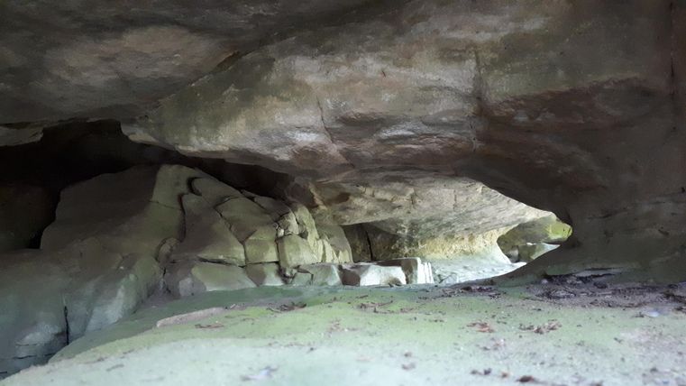 Eine natürliche Höhle mit großen, grauen Felsen. Der Boden ist sandig und es gibt wenig Licht.