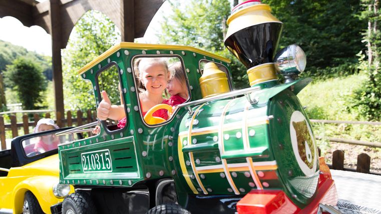 Ein Kind fährt fröhlich in einem bunten Miniaturzug. Die Sonne scheint, und die Umgebung ist grün und einladend.