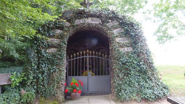 Eine mit Efeu bewachsene Lourdesgrotte mit einem Tor und Blumen davor.