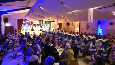 A festive event in a decorated room with many guests. People are sitting at tables and watching a performance on stage.
